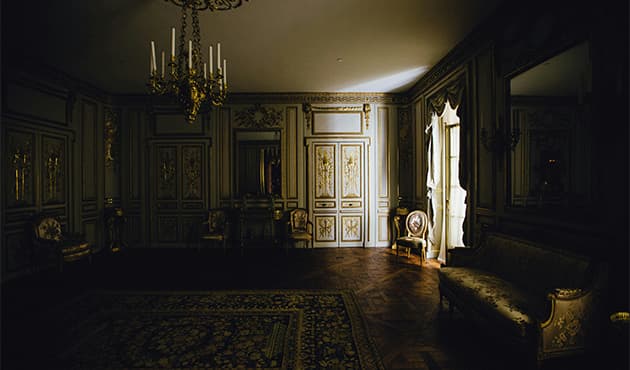 Dimly lit ornate room with gold doors, a chandelier, and a vintage chair next to a window.