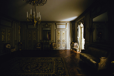 Dimly lit ornate room with gold doors, a chandelier, and a vintage chair next to a window.